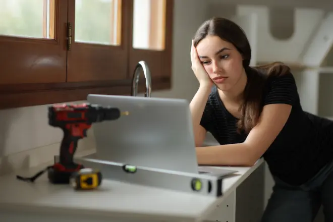 A person getting frustrated during a DIY kitchen remodel - LUA Construction A woman leans on a kitchen counter, looking at a laptop with a bored expression. A drill, a level, and a measuring tape are nearby.