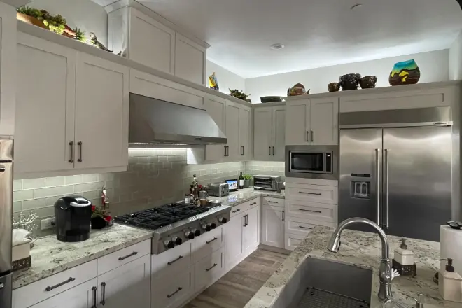 A remodeled kitchen by Lua Construction - LUA Construction Modern kitchen with white cabinets, stainless steel appliances, and marble countertops. A double-door fridge and a gas stove are present, with decorative items above the cabinets. DIY kitchen remodel cost